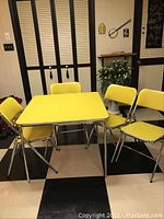 Full view of the yellow card table with four matching chairs arranged around it in room with black and white checkered floor.
