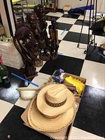 Photo showing five carved wooden statues arranged in a line on a black and white tiled floor, plus two straw hats and some table linens in a cardboard box.