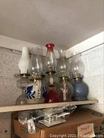 Eight vintage oil lamps displayed on a shelf showing a variety of base styles and chimney shapes.