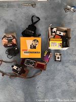 Overview of the vintage cameras and accessories including Kodak Brownie Hawkeye with case, Kodak Hawkeye Flash Outfit box, Kodak Tele Instamatic, Nikon One-Touch and other cameras and cases laid out on carpet.