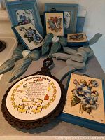 Floral themed artwork and plaques on kitchen counter including frames in blue and brown with attached cloth bows, and a kitchen prayer plaque in an ornate brown carved frame.