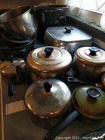 Photo of assorted metal pots with black handles and lids, black roaster and square roasting pan, and other cookware items stacked on countertop.