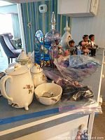 Overview of lot items including white ceramic teapot, sugar bowl, creamer, small porcelain bowl, purple candy dish, figurines and candles on a table.