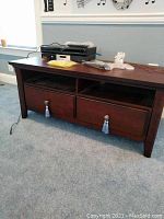 Front view of wood TV stand with two drawers, decorative tassel pulls, and open compartments, top surface holds electronics and accessories.