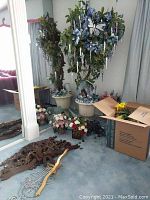 Two large faux floral small trees in pots with smaller floral arrangements around and a carved wood wall decor piece on floor.