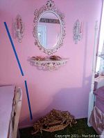 Room corner with oval floral mirror, matching wall shelf below, and two wall sconces on either side of mirror.