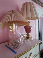 Photo of two table lamps on dresser, one lamp has a clear glass base with a cream pleated shade, the other has a pink ceramic and brass base with a beige shade.