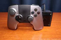 Silver and black Ouya game controller with analog sticks and colored buttons, placed on wooden surface with power adapter cable behind it.