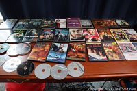 Photo showing a table with approximately 27 DVD cases arranged in rows, some loose discs stacked and spread on the table, titles include Harry Potter, Transformers, and others.