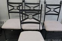 Four black metal dining chairs with light beige cushions arranged closely together, showing front and side views of frames and backrests.