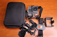 Safari WiFi camera, waterproof case, wrist remote, several mounts and the black carry case laid out on wooden surface.