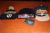 Photo showing all four items: three gaming-themed hats and one Zelda wallet placed on wooden surface