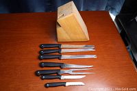Photo showing full set of six serrated kitchen knives with black handles arranged in front of a wooden knife block on a wood surface.