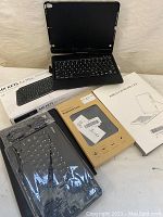 Photo showing multiple keyboards and keyboard cases including Logitech MX Keys for Mac keyboard in box, wireless keyboard case, and a rigid flip keyboard case with buttons visible inside.