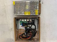 Top view of metal-cased power supply unit placed on top of brown cardboard box containing the open-frame power supply board and cables.