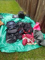Photo showing assortment of multiple sports and duffel bags in various colors and sizes on a green tarp outside near a wooden fence