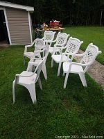 Seven white plastic lawn chairs on grass with different lattice and slatted back patterns, some showing dirt and wear.