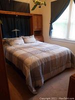 Queen size bed with gray-beige checkered bedding, showing the left side with a wooden cabinet under a shelf and cabinet doors.