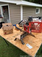 Miter saw and other tools set on wooden table outdoors with cardboard box and milk crate for storage