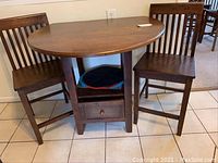 Round brown wooden dropleaf table flanked by two matching wooden chairs with storage drawer under seat.