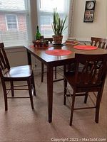 Wooden dining table with four chairs around it, placed in a corner with windows and some decorative items on the table removed for sale.