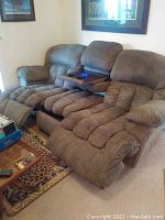 Full view of the brown reclining couch with pad segmented cushions, fold-down center tray with blue-lit control buttons, placed against a white wall with a framed picture above.