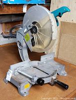 Side view of Makita miter saw showing 14-inch blade, blue handle, and cast iron base with quick-release mechanism and black adjustment knob.