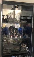 Shelf display of various glass, crystal, pewter items including decanters, pewter pitcher, blue glass items, plates, and serving trays.