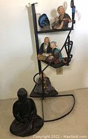 Three Hakata dolls placed on a black metal corner shelf with a black Buddha statuette on floor in front.