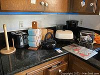 Countertop photo displaying all items in the lot including Oster toaster, teapot, coffee maker, skillet, wooden containers, paper towel holder, cups, and cutting board.