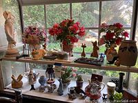 Wide view of decorative items on glass shelves featuring faux flowers, cat figurines, and ceramic collectibles.