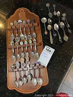 Full view of collection of 19 assorted souvenir spoons on a wooden rack, showing mixed designs and colors.