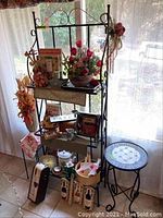 Baker's rack with three shelves, decorated with floral arrangements and various items, positioned near a window with a sheer curtain. Small round mosaic tile top table placed to the right of the rack.