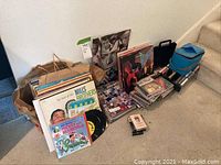 Assortment of vinyl records, CDs, and audio tapes arranged on floor showing several album covers and stacks.