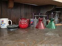 Photo of glassware including purple tinted stemmed glasses, red and green colored inverted cone glasses, white mug with bird cartoon, red textured glass jars, black scalloped edge ashtray or candle holder, and candle.