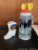Blue and white ceramic beer stein with Praha and a small white boot-shaped ceramic vase with scenic decal on black surface, against wood background