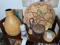 Photo showing large wicker vase, woven tray, two doll figures, ceramic bell, lidded basket, small round basket, coasters, and sand art bottle on a chair.