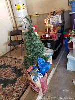 Artificial Christmas tree, cardboard bag with Christmas lights box, gold reindeer figurine, and stacked folding tables in storage area.