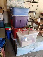 Stack of 10 plastic storage containers in various colors with holiday decorations inside, topped with a green metal tree stand and red-white garland wrapped around it.