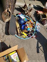 Photo of various toys and recreation items including skateboard, bowling ball, baseball bats, hand pump, hula hoop, and assorted small toys laid out on pavement.