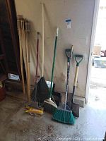Full view of garden tools and tiki torches leaning against wall showing variety of handles and bristles