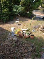 Wide outdoor photo showing group of garden items including pots, statues, and a white birdbath in a yard covered with dry leaves and branches.