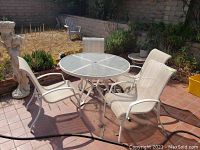 Patio glass top round table with 4 white metal framed mesh chairs arranged around it on a tiled patio with some garden elements and pots in the background.