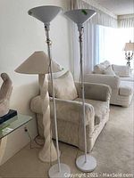 Two white metal floor lamps with wide metal shades standing side by side in a living room.