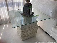 Full view of side table showing square clear glass top and faux marble textured base next to a white couch.