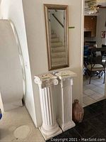 Two classical column pedestals with decorative capitals next to a wall mirror against wall, decorative vessel on floor