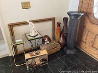 Wide view of the assortment showing two large picture frames, two column pedestals, three tall vases, a footstool with a plush dog, and a small metal and glass side table.