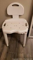 White plastic bath chair front view, showing backrest with handle and perforated seat with legs on hardwood floor.
