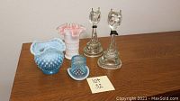 Photo showing entire lot including two Hemingray cat glass insulators, blue hobnail candy dish, pink and white swirled ruffled vase, and blue hobnail teacup with saucer on wooden table.