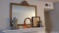 Wide shot of mantel showing the gold framed mirror, angel statue, boy with bunny statue, framed art print, and small oval frame